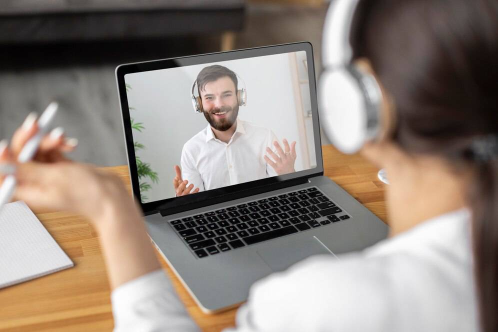 A person engages in a video call on a laptop, wearing headphones and gesturing while speaking, with greenery in the background. 