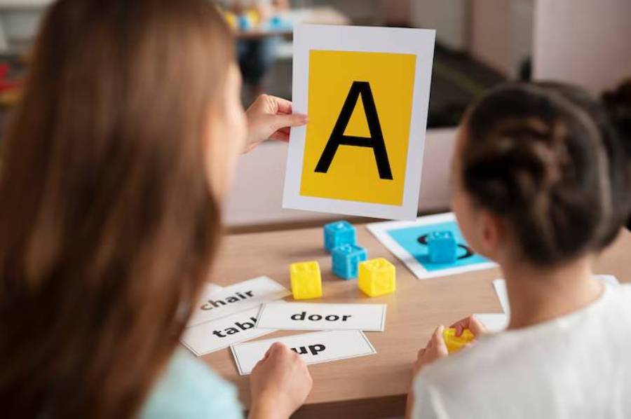  Two children learning letters, with one holding a bright yellow card displaying the letter A while colorful blocks and word cards are on the table.