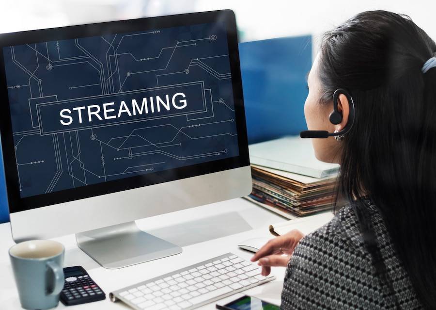 A woman with a headset sits at a desk, focused on a computer screen displaying the word STREAMING in bold text.