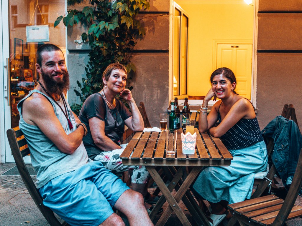  A group of three people sit around a wooden table, drinks in hand, with a cozy outdoor atmosphere and warm lighting in the background.