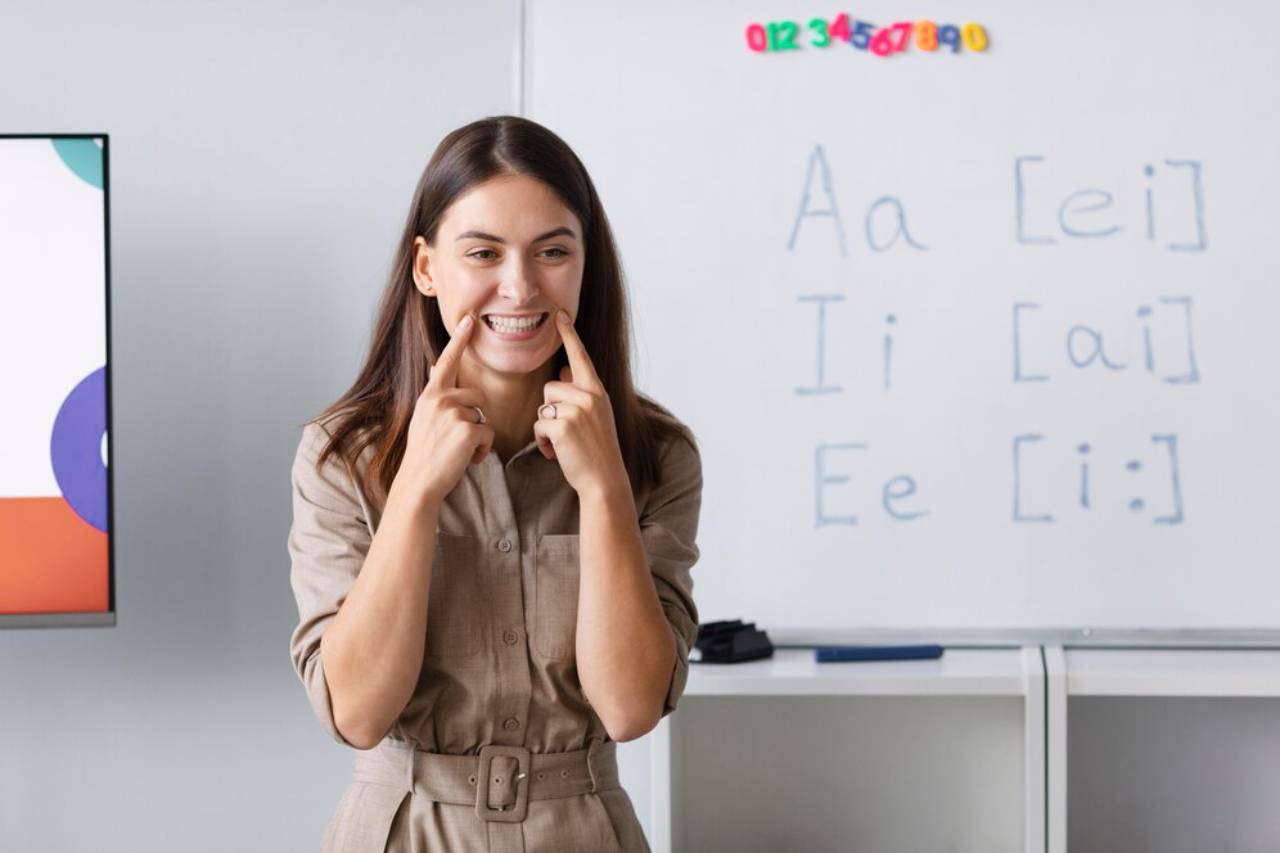 A person in a beige outfit stands in front of a whiteboard with letters and phonetic symbols, engaged in a classroom setting.
