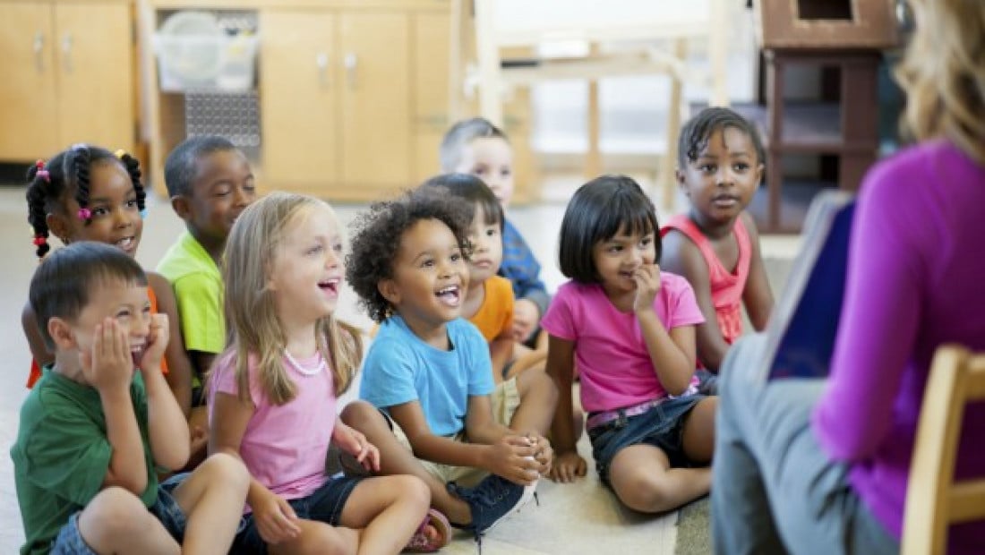 A group of diverse children sit attentively on the floor in a classroom, listening to an adult speaking in front of them.