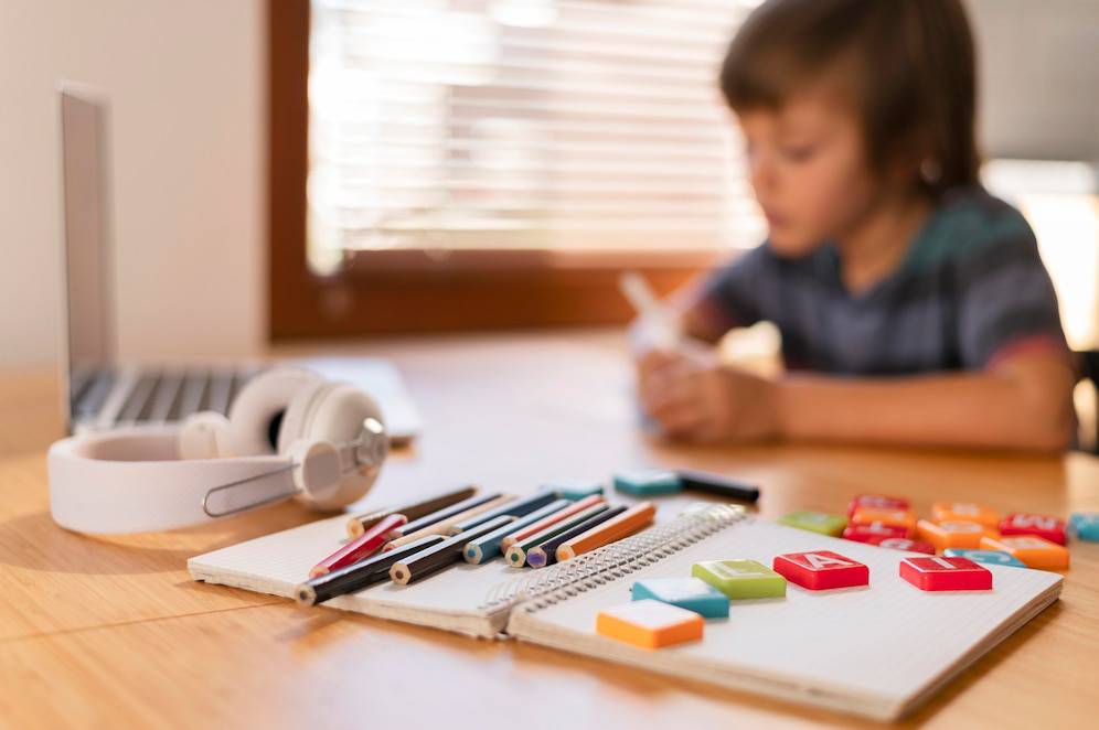  A wooden table featuring a laptop, headphones, colorful pens, and letter tiles arranged on a notepad. Natural light from a window illuminates the scene.