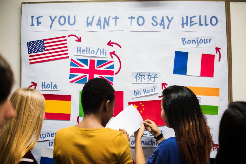 A diverse group of people studies a multilingual Hello poster, featuring flags and greetings in various languages. 
