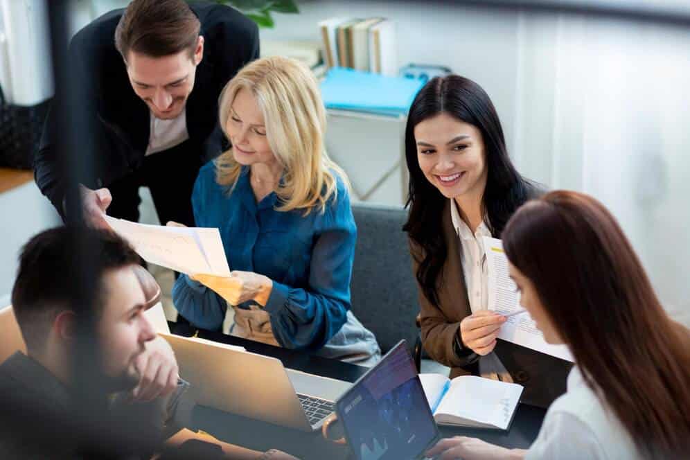A group of professionals engages in a collaborative meeting, reviewing documents and sharing ideas around a table with laptops and notepads.