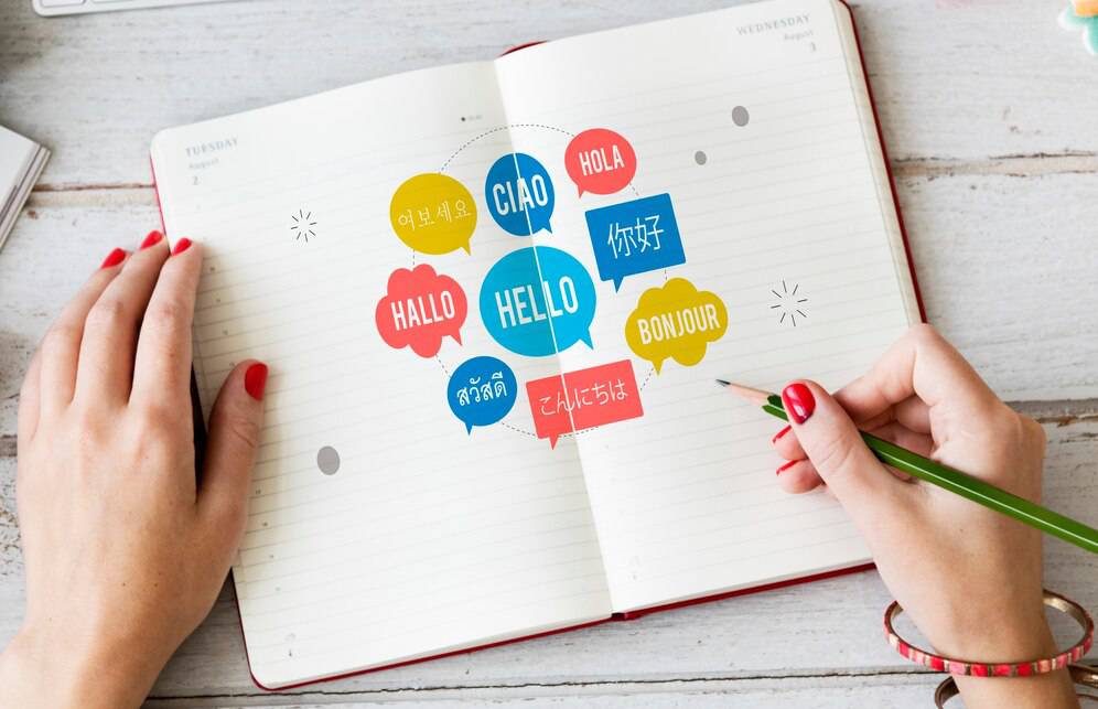 A hand with red nails writes in a notebook surrounded by colorful speech bubbles greeting in multiple languages. 