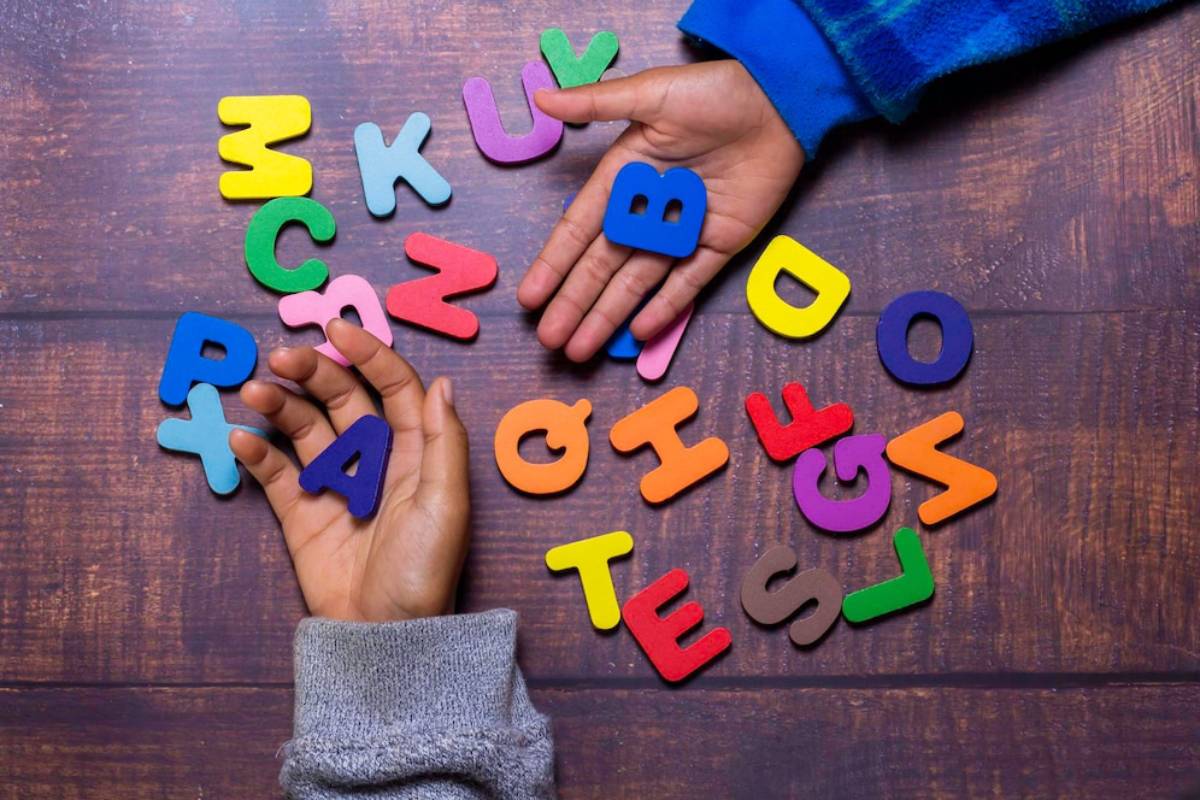 Two hands hold and arrange colorful wooden alphabet letters scattered on a wooden surface. 