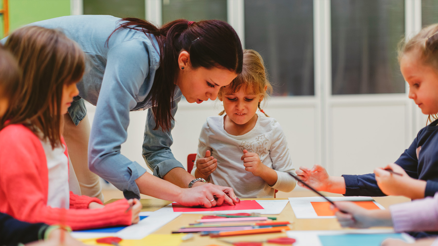 A teacher guides children in an art class, surrounded by colorful paper and art supplies on a table.