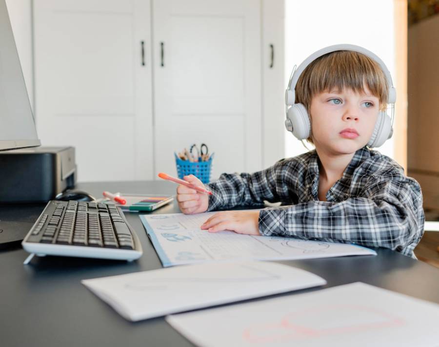  A child wearing headphones sits at a desk with a computer, holding a colored pencil, surrounded by worksheets and stationery.