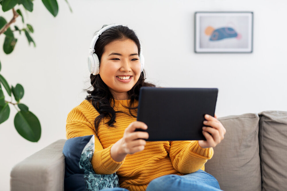 A person in a cozy living room wears headphones and holds a tablet, sitting comfortably on a couch with a decorative pillow. 