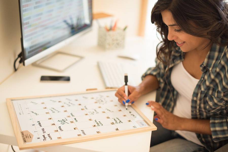 A person writing on a calendar planner at a desk, with a computer and smartphone in the background, surrounded by office supplies. 