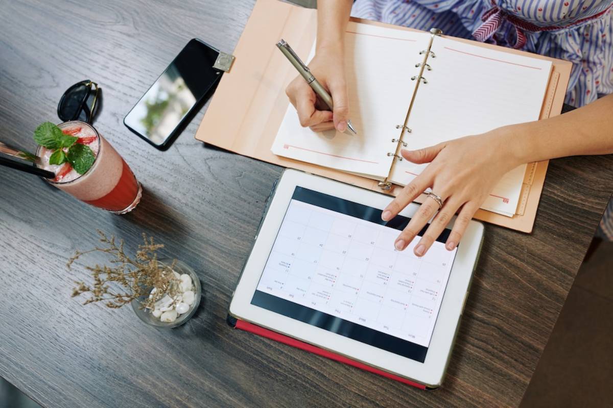 A person writes in a planner while using a tablet to view a calendar, with a drink and phone on a wooden table. 