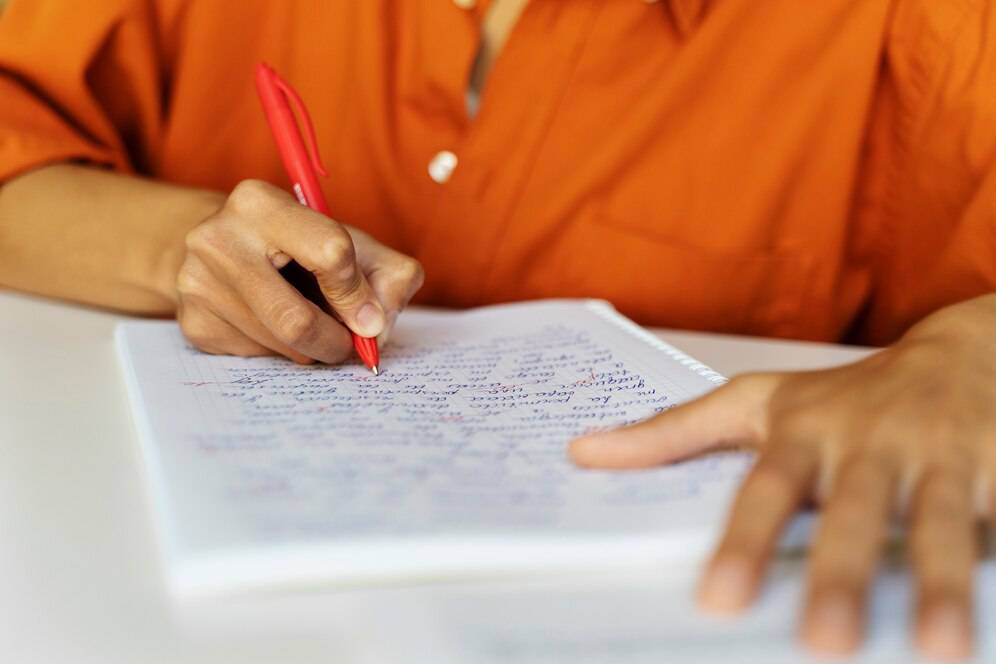  A close-up of a person in an orange shirt writing in a notebook with a red pen, surrounded by handwritten notes.