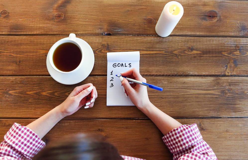  A person in a checkered shirt writes down goals in a notebook, with a cup of tea and a candle on a wooden table.