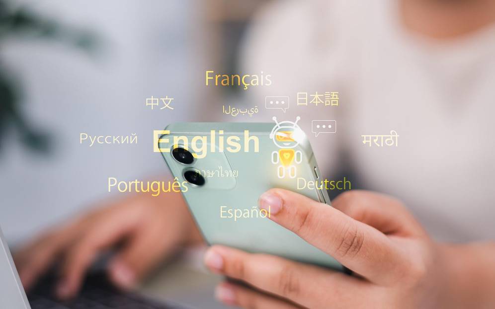  A person holds a smartphone displaying various languages, including English, while working on a laptop in a modern setting.