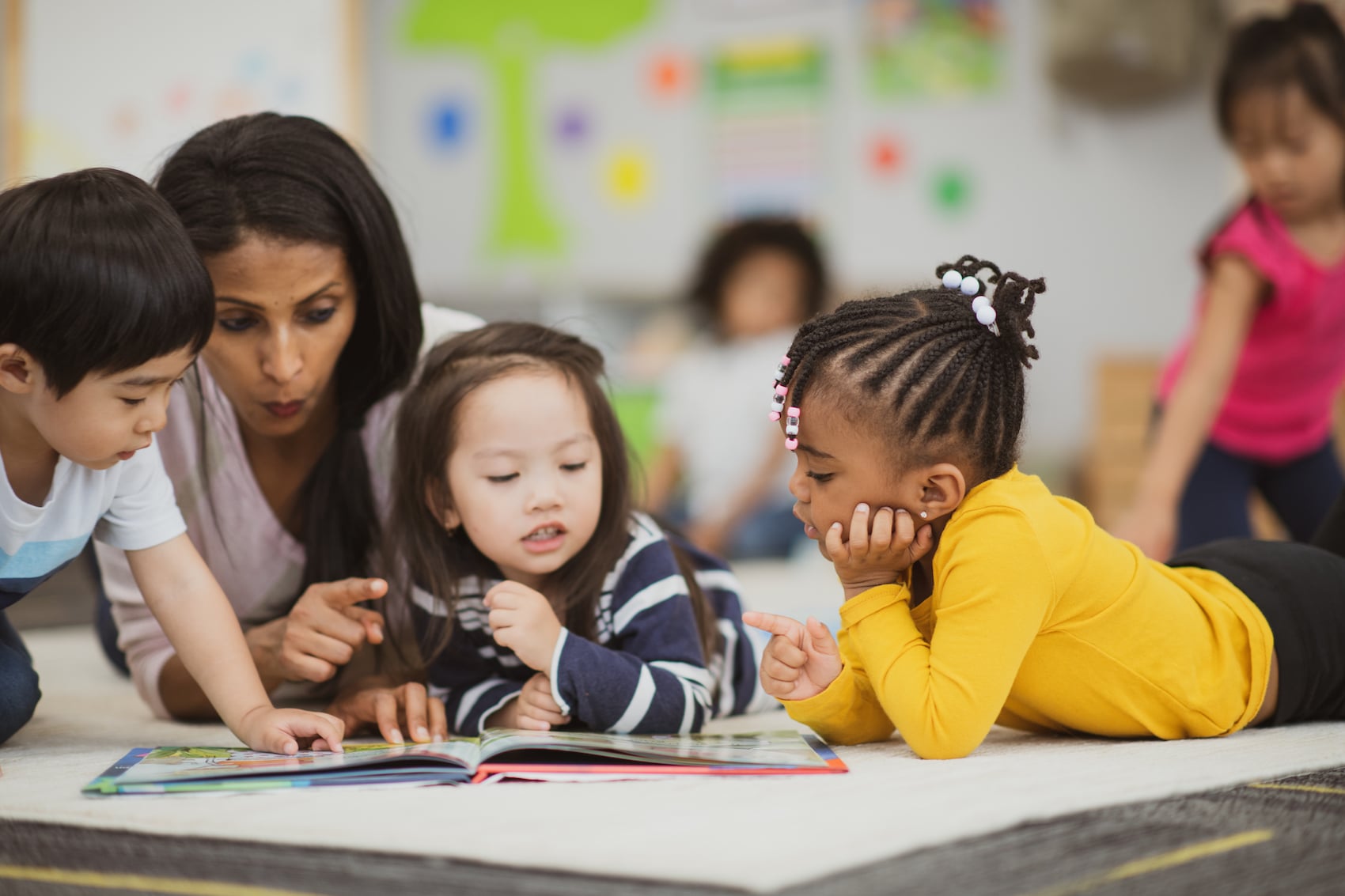 A diverse group of children engaging with a picture book on the floor, surrounded by educational materials in a bright classroom.