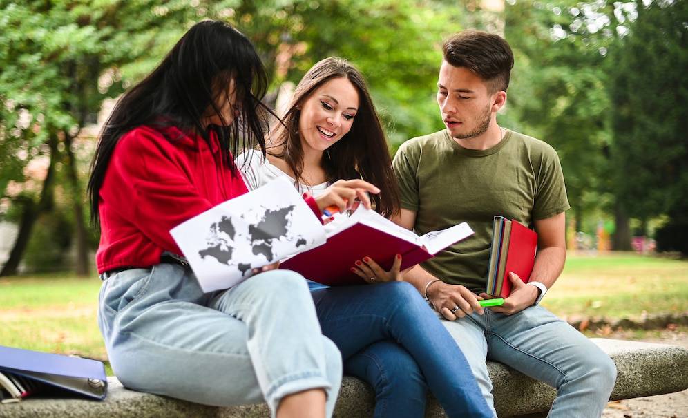  Three young adults sit outdoors, engaged in a study discussion with books and a map in hand, surrounded by greenery.