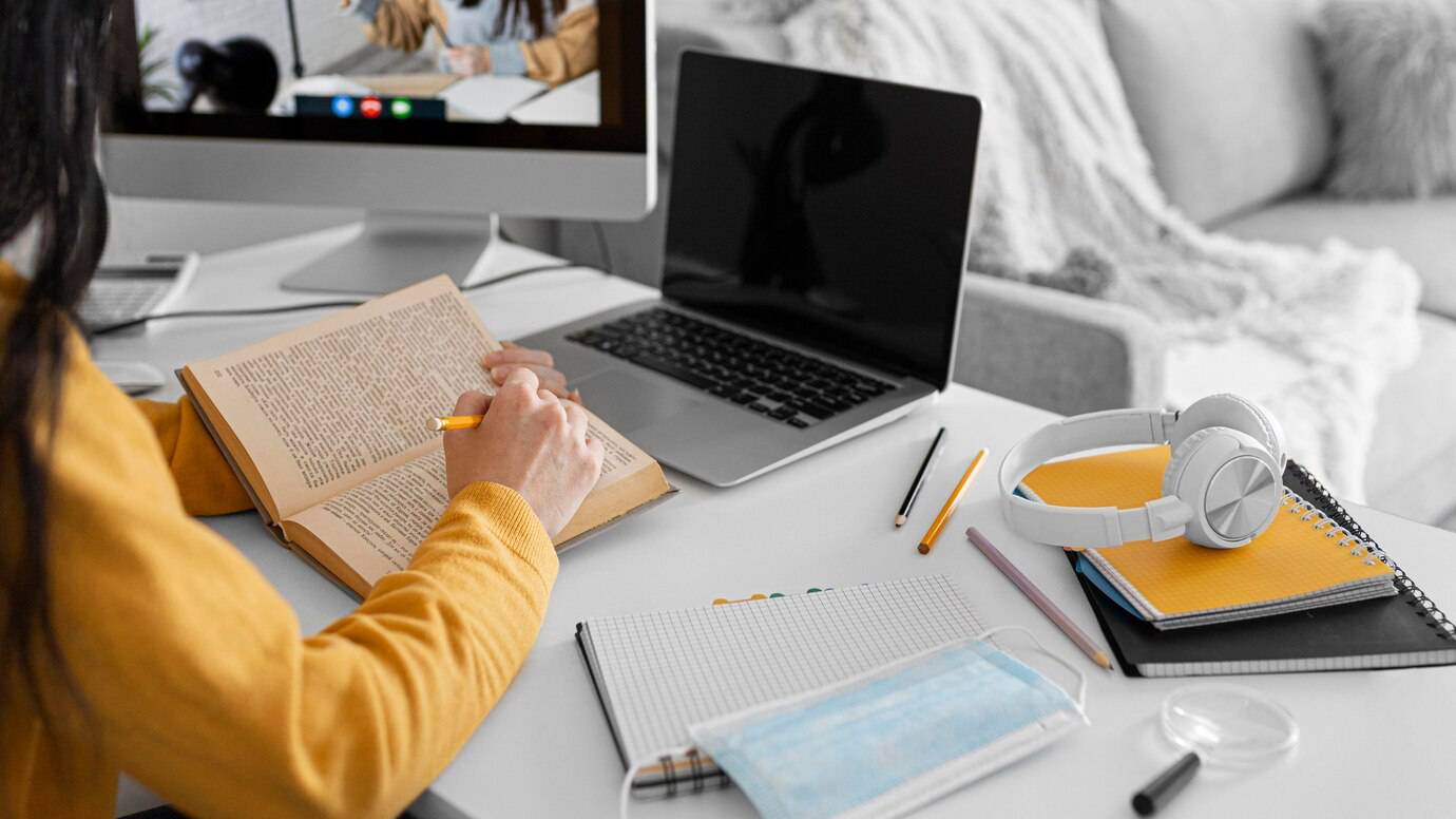 A person in a yellow sweater studies from a book, surrounded by a laptop, headphones, notebooks, and a face mask on a desk.
