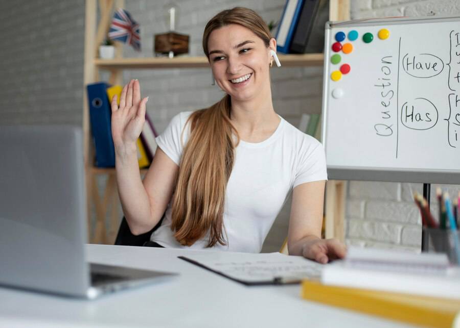 A person with long hair in a white shirt is sitting at a desk, gesturing with their hand, with a laptop and a whiteboard in the background. 