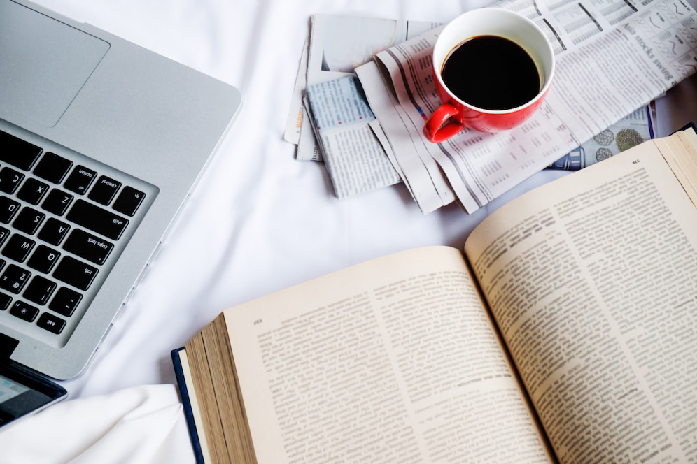 A laptop, an open book, and a red coffee cup sit on a white surface alongside scattered newspapers. 