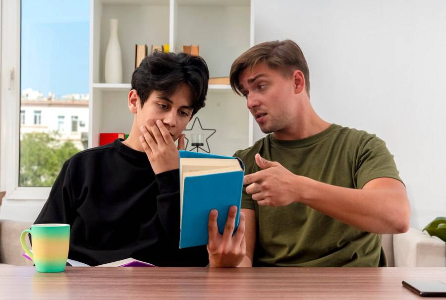  Two individuals engage in a discussion over a blue book, seated at a table with a colorful mug nearby, in a bright, modern room.