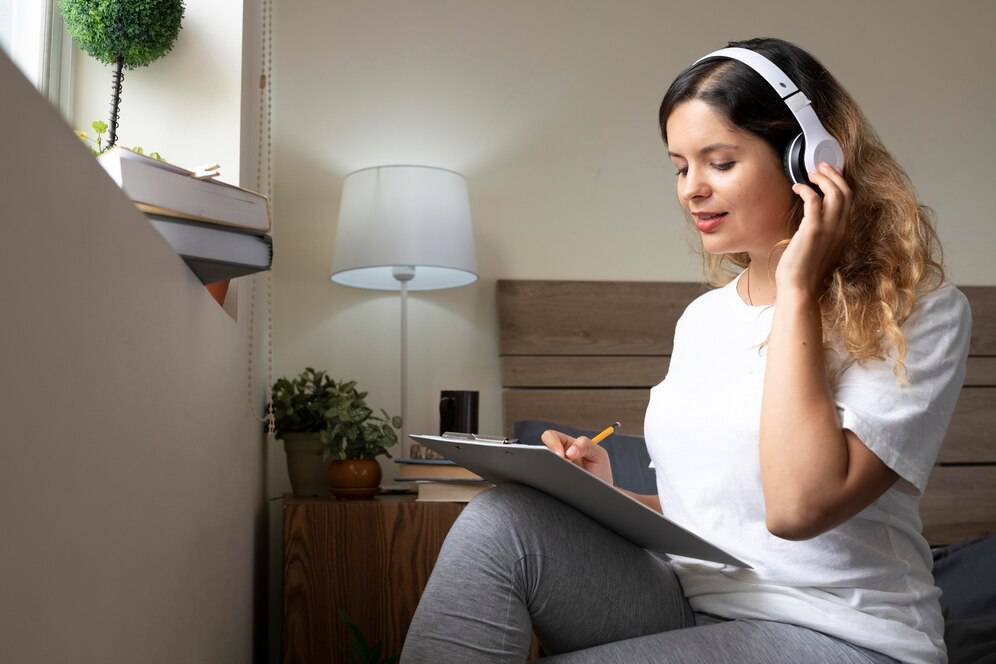 A person wearing headphones sits on a bed, taking notes on a clipboard in a cozy, well-lit room with plants and a lamp.