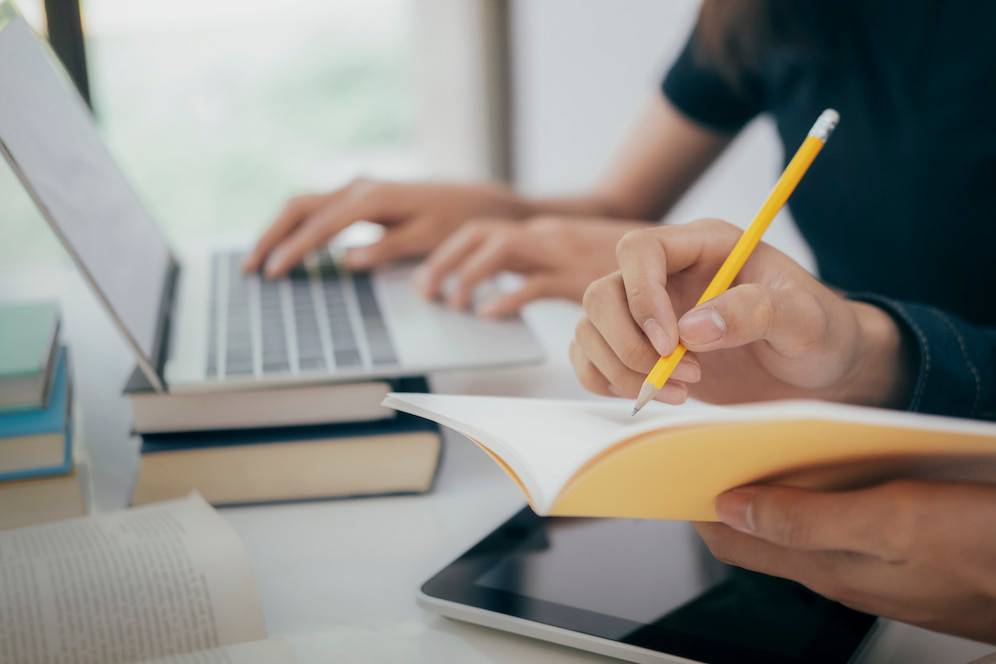 A person writes in a yellow notebook with a pencil while another types on a laptop, surrounded by books and a tablet on the desk.