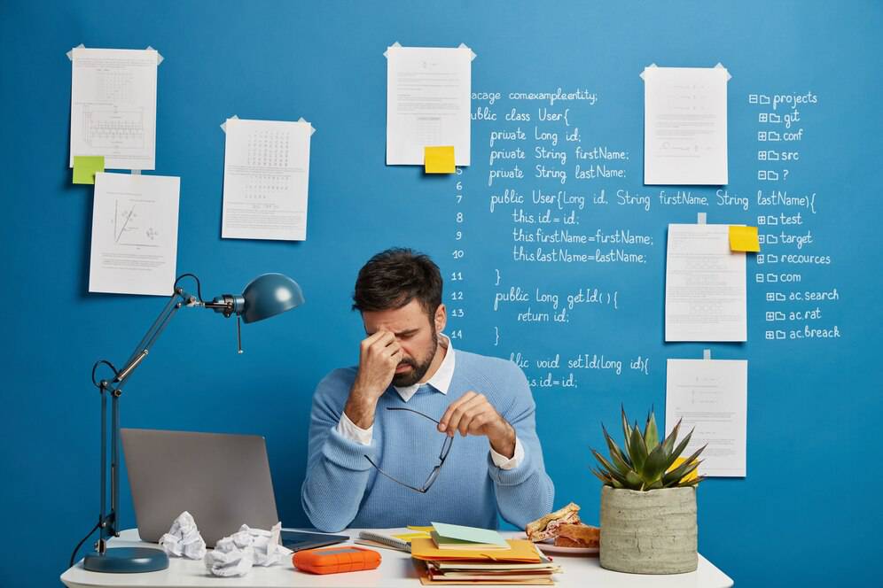 A person sits at a desk with a laptop, surrounded by notes and diagrams on a blue wall, and a plant beside them.