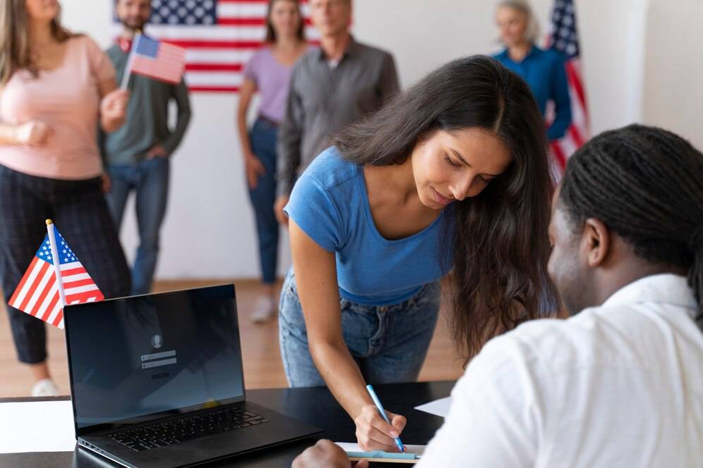 A person is signing a document at a table, with an American flag on the desk and several individuals celebrating in the background. 