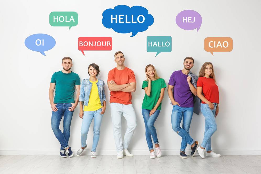 A group of six people stands against a white wall, displaying speech bubbles with greetings in various languages. 