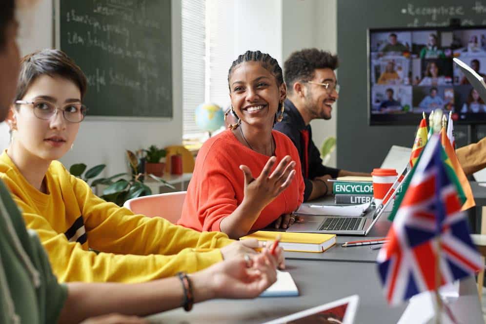 A group of students engaged in a discussion at a table, with laptops and notebooks, and a screen displaying a video conference.