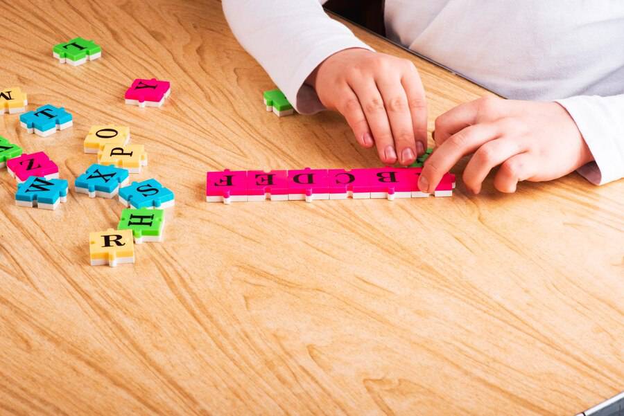 A child’s hands manipulate colorful letter tiles on a wooden table, forming the word EDGE with pink tiles. 