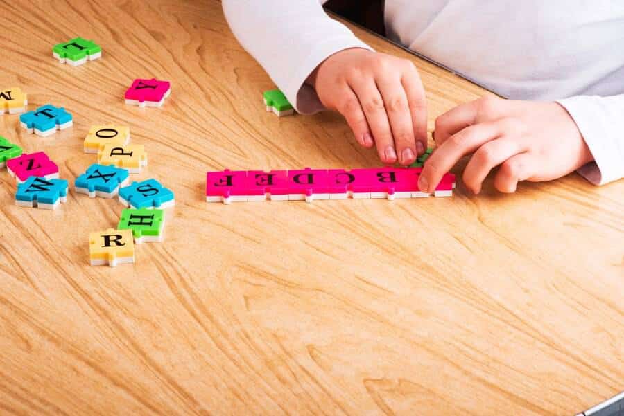 A child’s hands manipulate colorful letter tiles on a wooden table, forming the word EDGE with pink tiles.