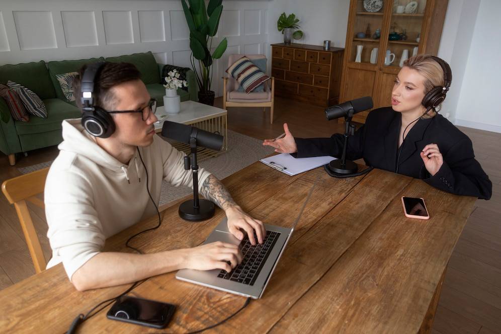 Two podcast hosts engage in conversation at a wooden table, with microphones and a laptop, set in a cozy living room.