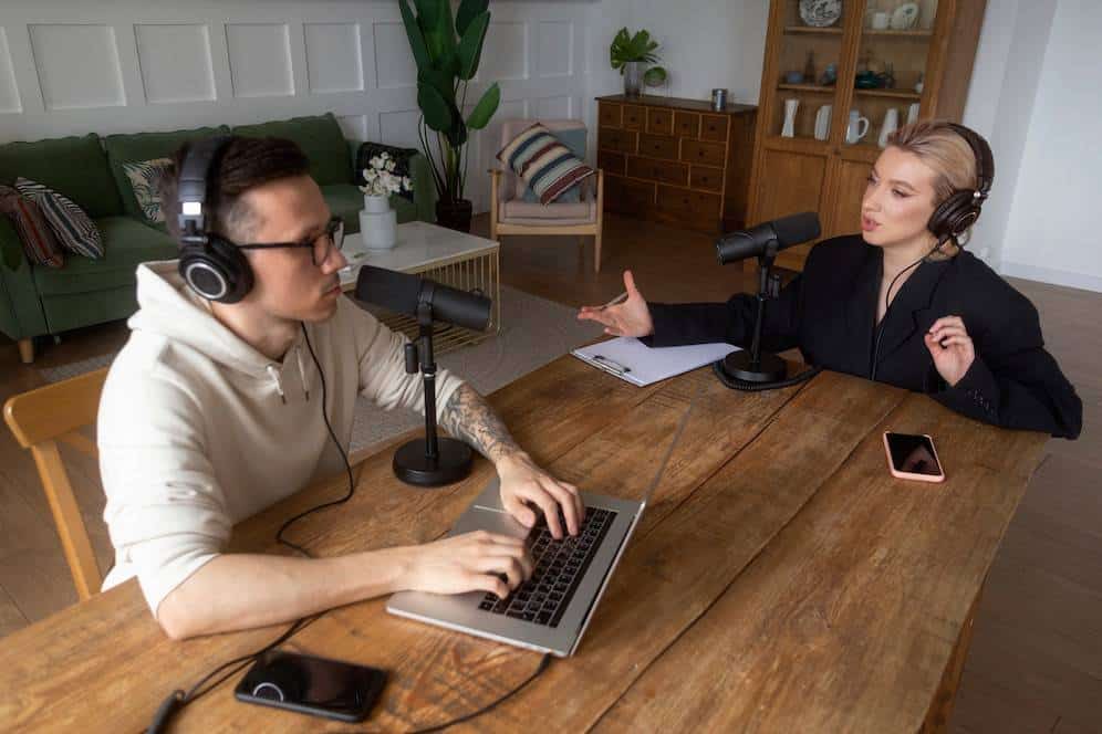 Two podcast hosts engage in conversation at a wooden table, with microphones and a laptop, set in a cozy living room.