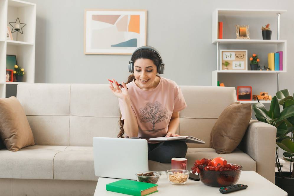 A person with long hair wearing headphones sits on a couch, engaged in a virtual session, with snacks and a laptop on a table.
