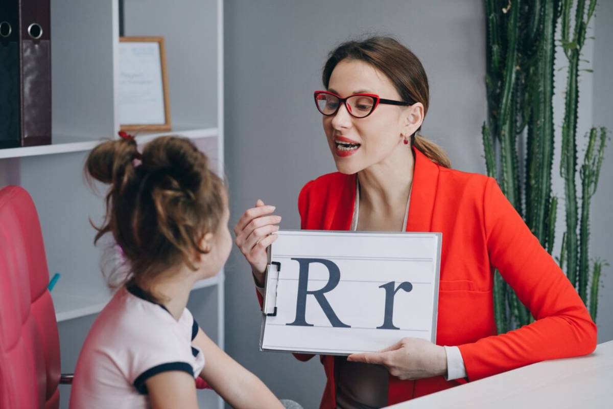 A woman in a red blazer holds a sign displaying the letter R while engaging with a young girl in a classroom setting.
