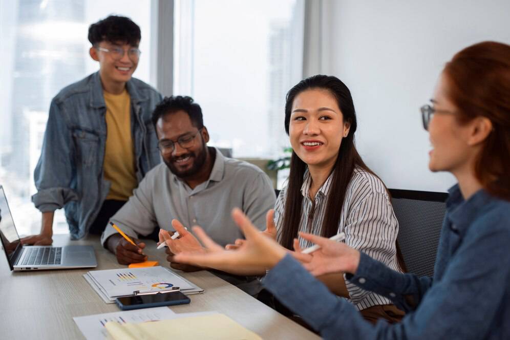 A diverse group of professionals engaged in a discussion around a table with a laptop and documents in a bright office environment. 