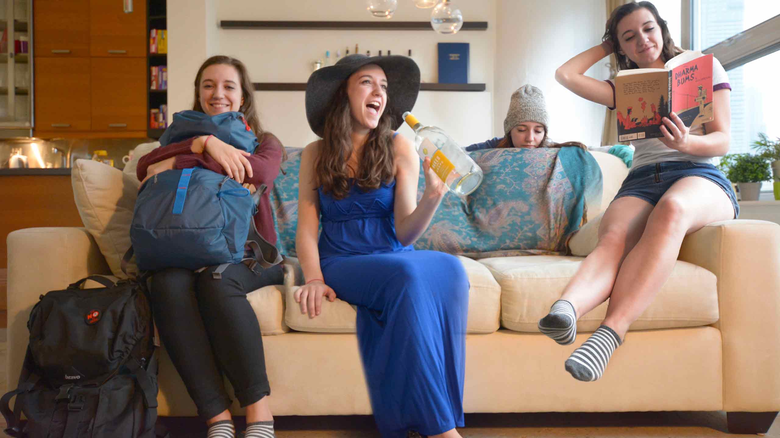  A group of friends relaxing on a couch, with one holding a drink, another reading a book, surrounded by backpacks and colorful decor.