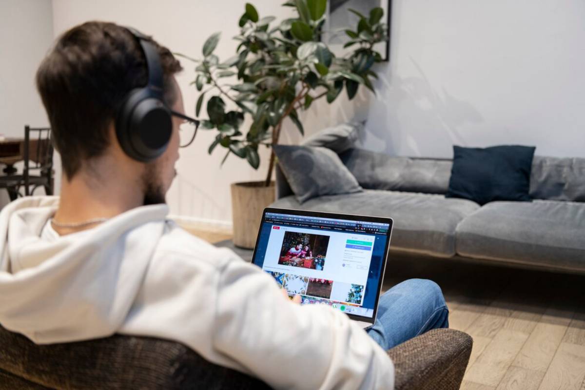 A man in headphones sits on a couch, focused on his laptop, with a potted plant nearby and a cozy living room in the background.