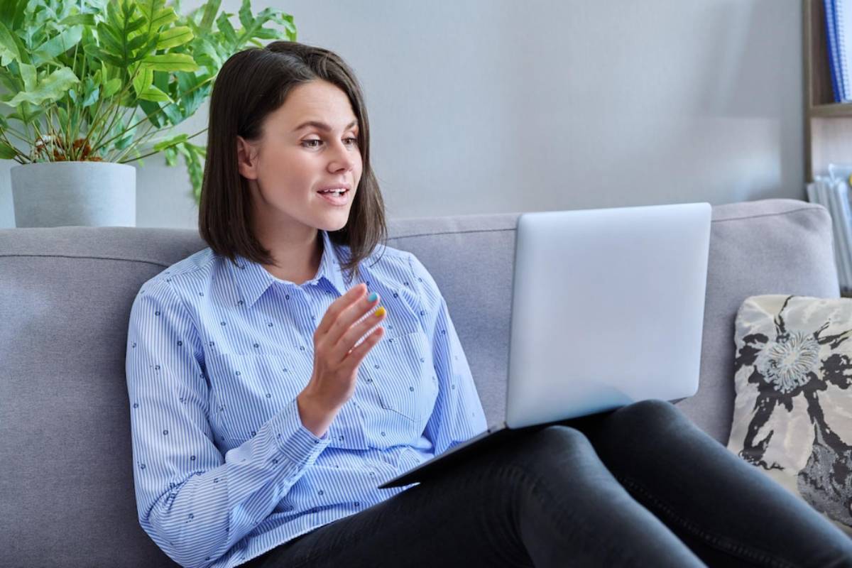  A person in a blue striped shirt sits on a sofa, gesturing while using a laptop. Green plants are visible in the background.