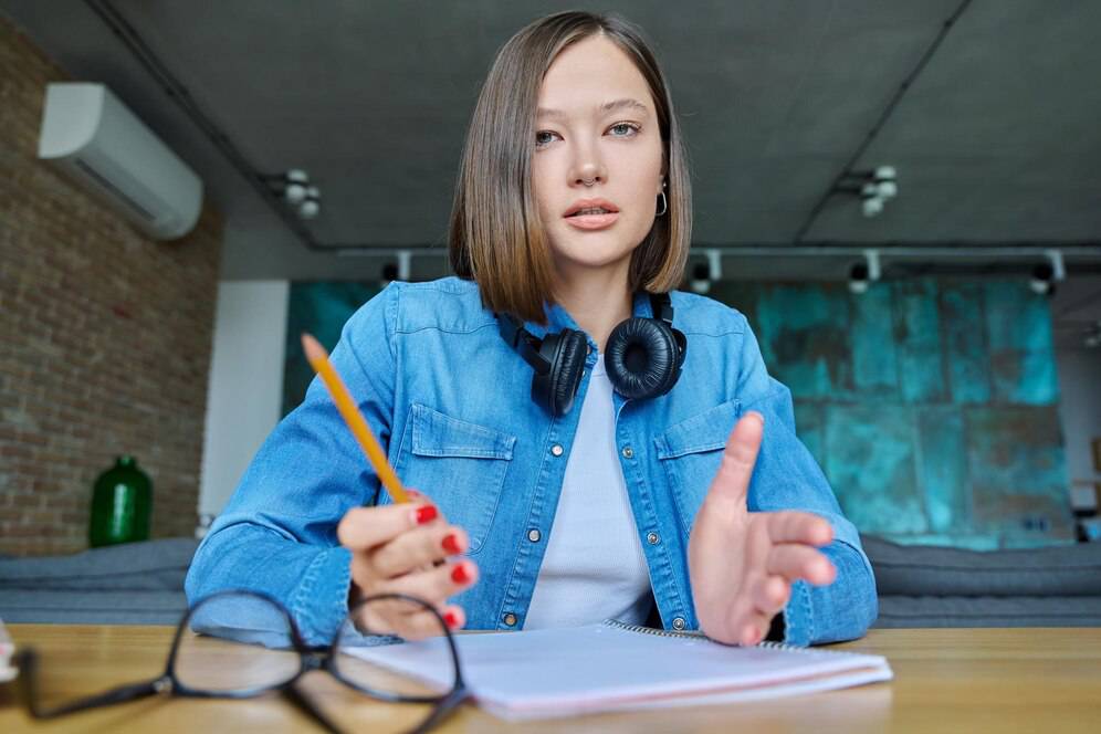 A person wearing a denim jacket and headphones sits at a table, holding a pencil, with an open notebook and glasses nearby. 