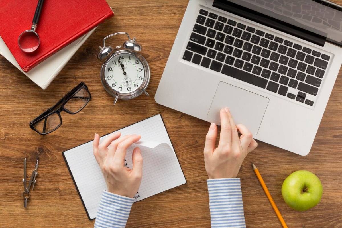  A top-down view of a workspace featuring a laptop, notebook, clock, glasses, a pencil, an apple, and a magnifying glass.