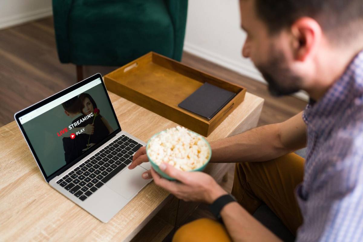  A person holds a bowl of popcorn while watching a live stream on a laptop, sitting at a cozy wooden table.