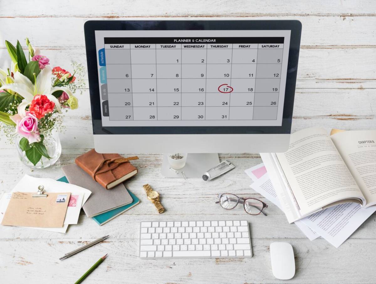 A desktop computer displays a calendar, with a circled date, surrounded by flowers, notebooks, glasses, and stationery.