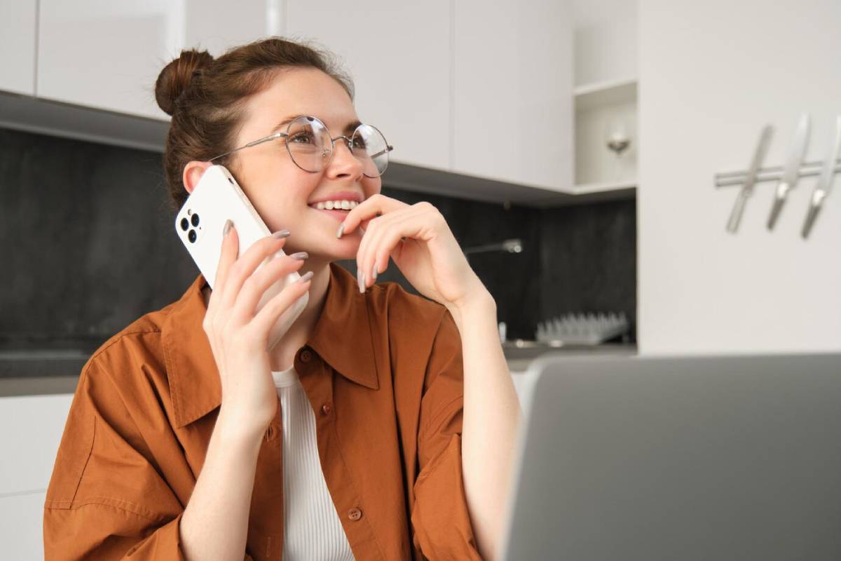 A woman in a brown jacket is talking on her phone, with a laptop open in front of her in a modern kitchen setting. 